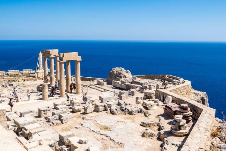 View od the remaining columns and pediments on the top of the Acropolis of Lindos with the dark blue Aegean Sean far below