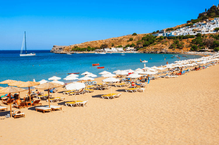 Photo of sunny day with sun loungers and umbrellas at Lindos Beach on Lindos Bay, Rhodes